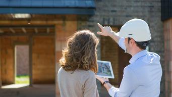 Mann mit Bauhelm zeigt auf ein Tablet, während eine Frau in einem Baustelleninneren zuschaut.